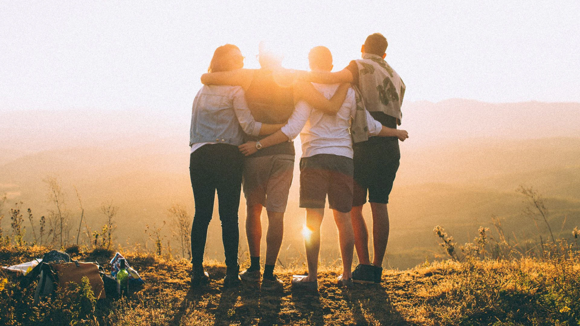 Vier junge Personen umarmen sich und sehen sich auf einem Aussichtspunkt den Sonnenuntergang an.